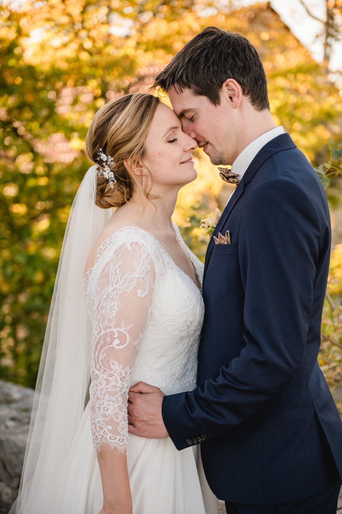 Braut und Bräutigam umarmen sich zärtlich im Freien, im Hintergrund herbstliches Laub. Die Braut trägt ein Spitzenkleid und einen Schleier, der Bräutigam einen marineblauen Anzug.