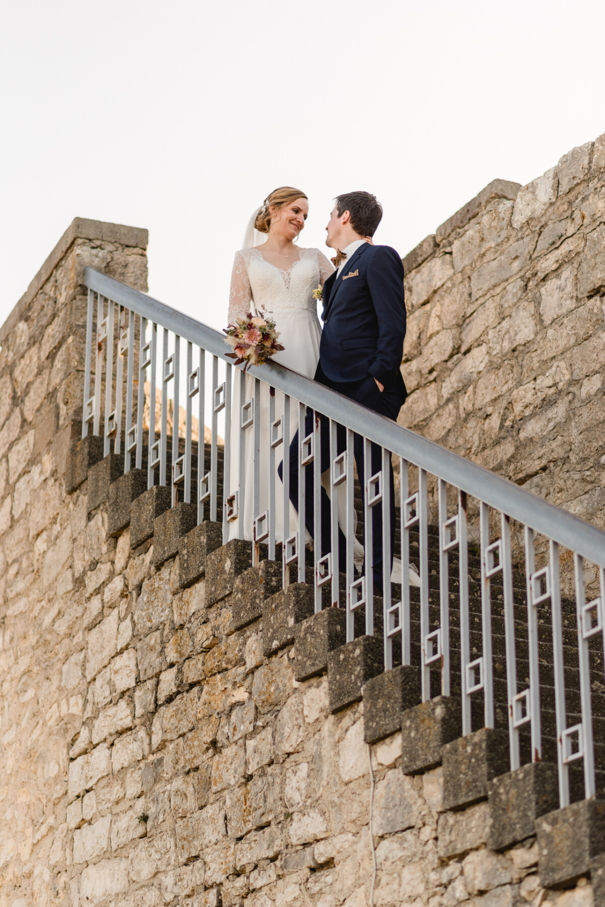 christian-lorenz.net Braut und Bräutigam stehen gemeinsam auf einer Steintreppe, in einem weißen Kleid und einem dunklen Anzug, mit einem Blumenstrauß in der Hand, vor der Kulisse einer alten Steinmauer.