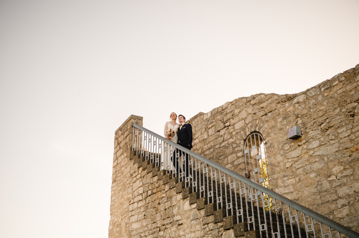 Braut und Bräutigam stehen gemeinsam auf einer Steintreppe im Freien, im Hintergrund ein klarer Himmel.