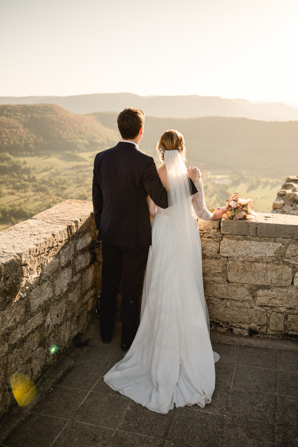 Braut und Bräutigam stehen auf einer Steinterrasse und blicken während des Sonnenuntergangs auf eine malerische Landschaft.