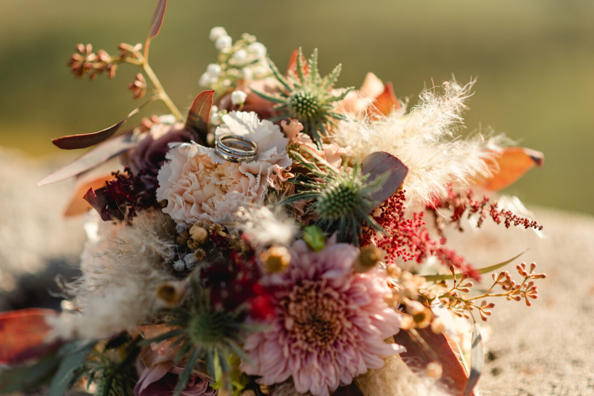 christian-lorenz.net Ein Strauß mit verschiedenen Blumen und Grünpflanzen, mit zwei Ringen zwischen den Blüten, platziert auf einer Steinoberfläche.
