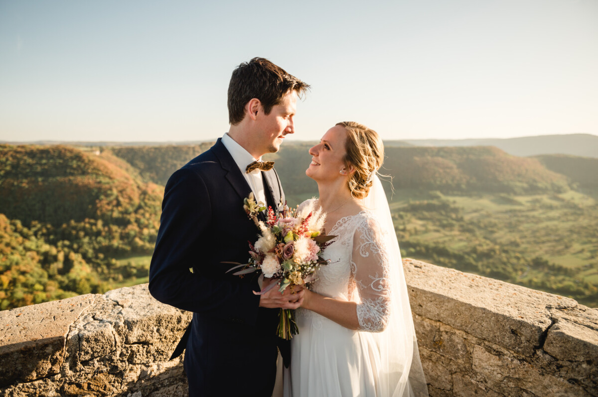 Ein Paar im Hochzeitskleid steht auf einer Steinterrasse mit Blick auf ein malerisches Tal, lächelt sich an und hält einen Blumenstrauß in der Hand.