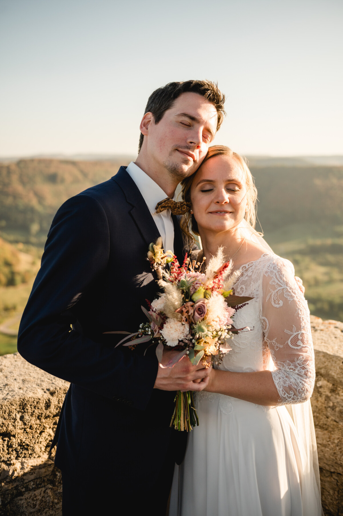 Braut und Bräutigam umarmen sich lächelnd mit geschlossenen Augen. Die Braut hält einen Blumenstrauß in der Hand. Sie stehen im Freien vor einer sanften Hügelkulisse unter klarem Himmel.