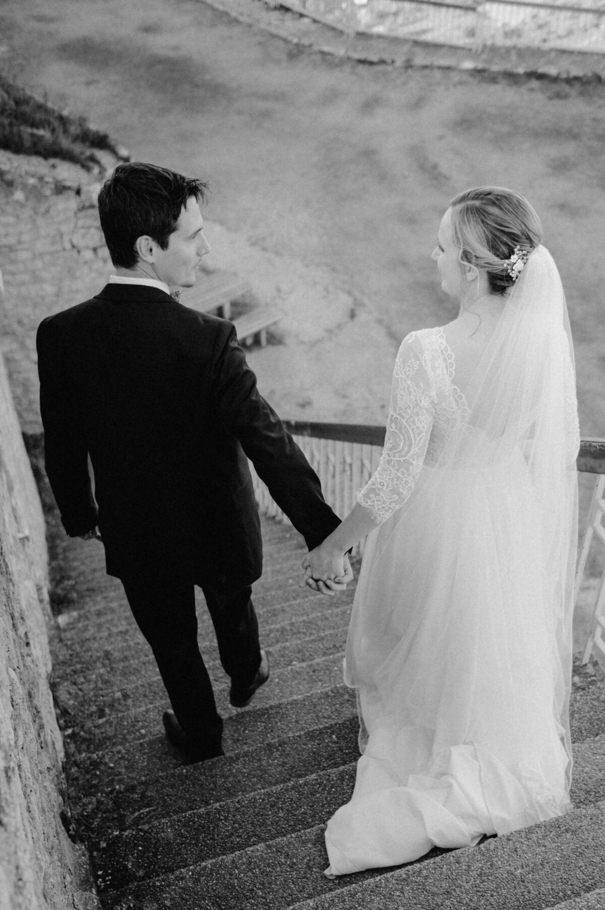 Braut und Bräutigam gehen Händchen haltend die Treppe hinunter. Die Braut trägt einen langen Schleier und ein Spitzenkleid, der Bräutigam einen dunklen Anzug. Die Szene spielt im Freien vor einem felsigen Hintergrund.
