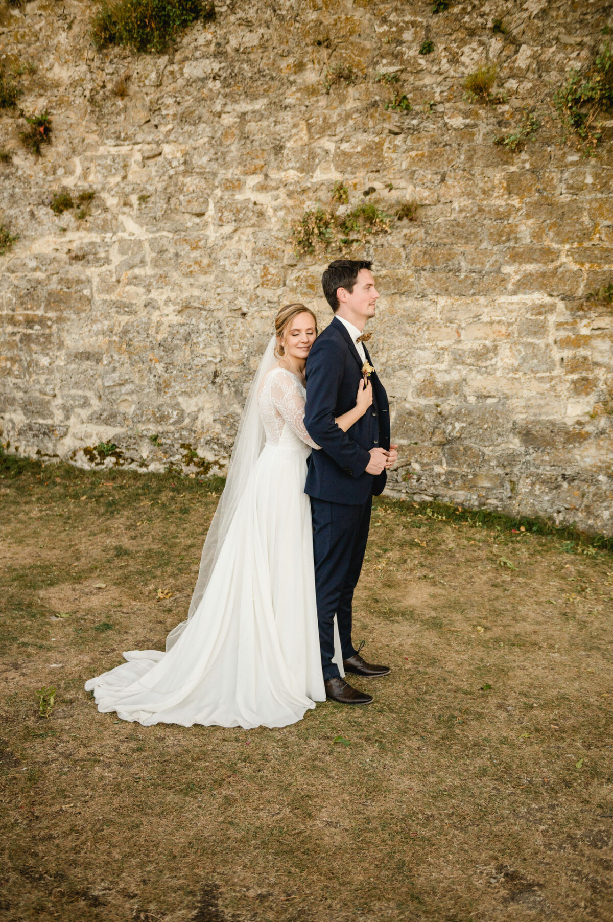 Eine Braut in einem weißen Kleid umarmt einen Bräutigam in einem marineblauen Anzug von hinten, der auf dem Gras vor einer Steinmauer steht.