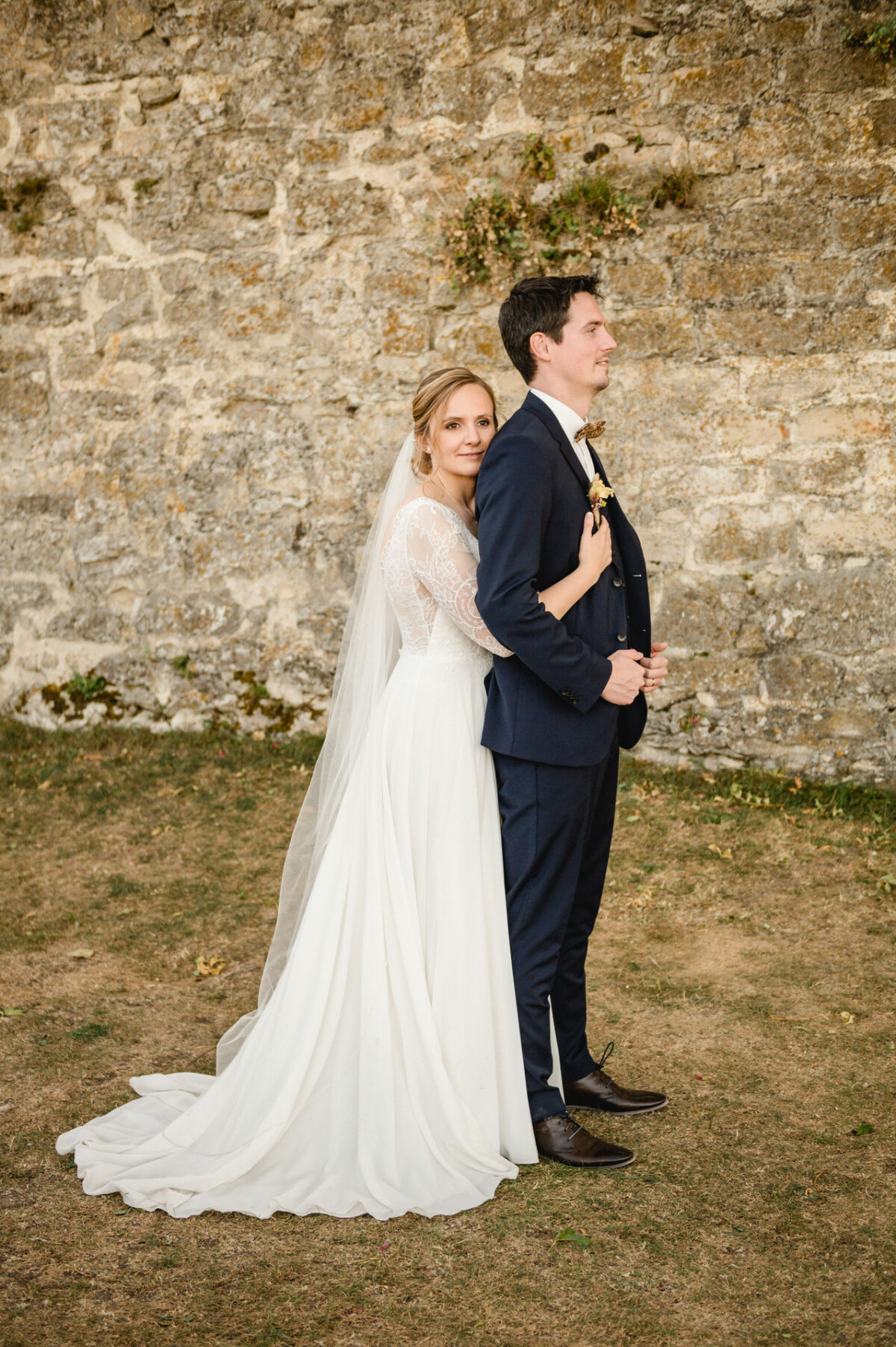Braut im weißen Kleid und Bräutigam im dunklen Anzug stehen draußen zusammen vor einer Steinmauer.
