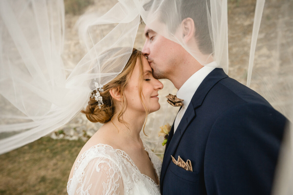 Braut und Bräutigam umarmen sich unter einem Schleier. Der Bräutigam küsst die Braut auf die Stirn. Beide wirken fröhlich und ruhig.