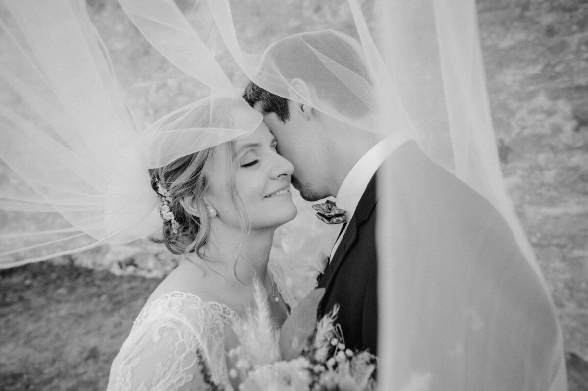 Braut und Bräutigam unter einem Schleier, eng beieinander mit geschlossenen Augen, in einem zärtlichen Moment. Schwarz-Weiß-Foto.