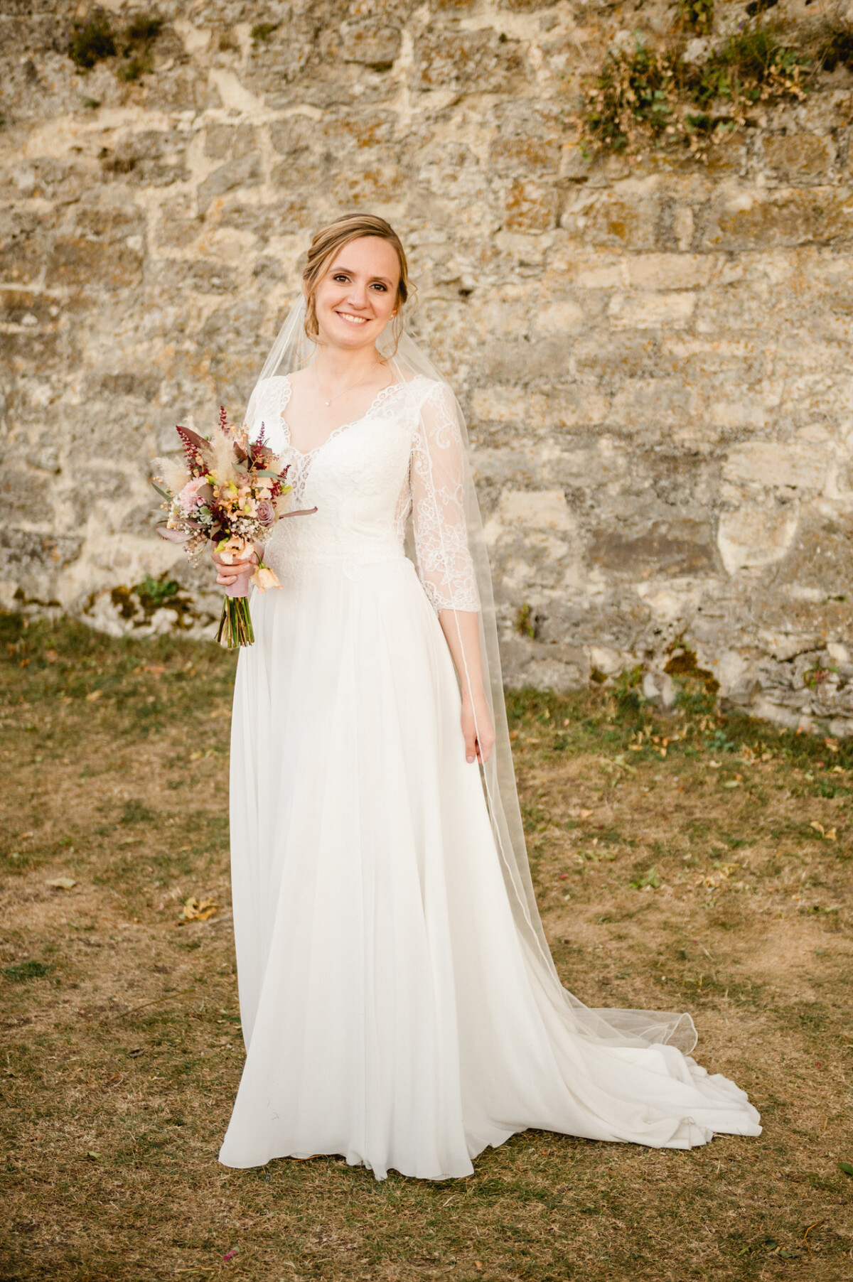 Braut in einem weißen Kleid hält einen Blumenstrauß, steht lächelnd auf dem Gras vor einer Steinmauer.