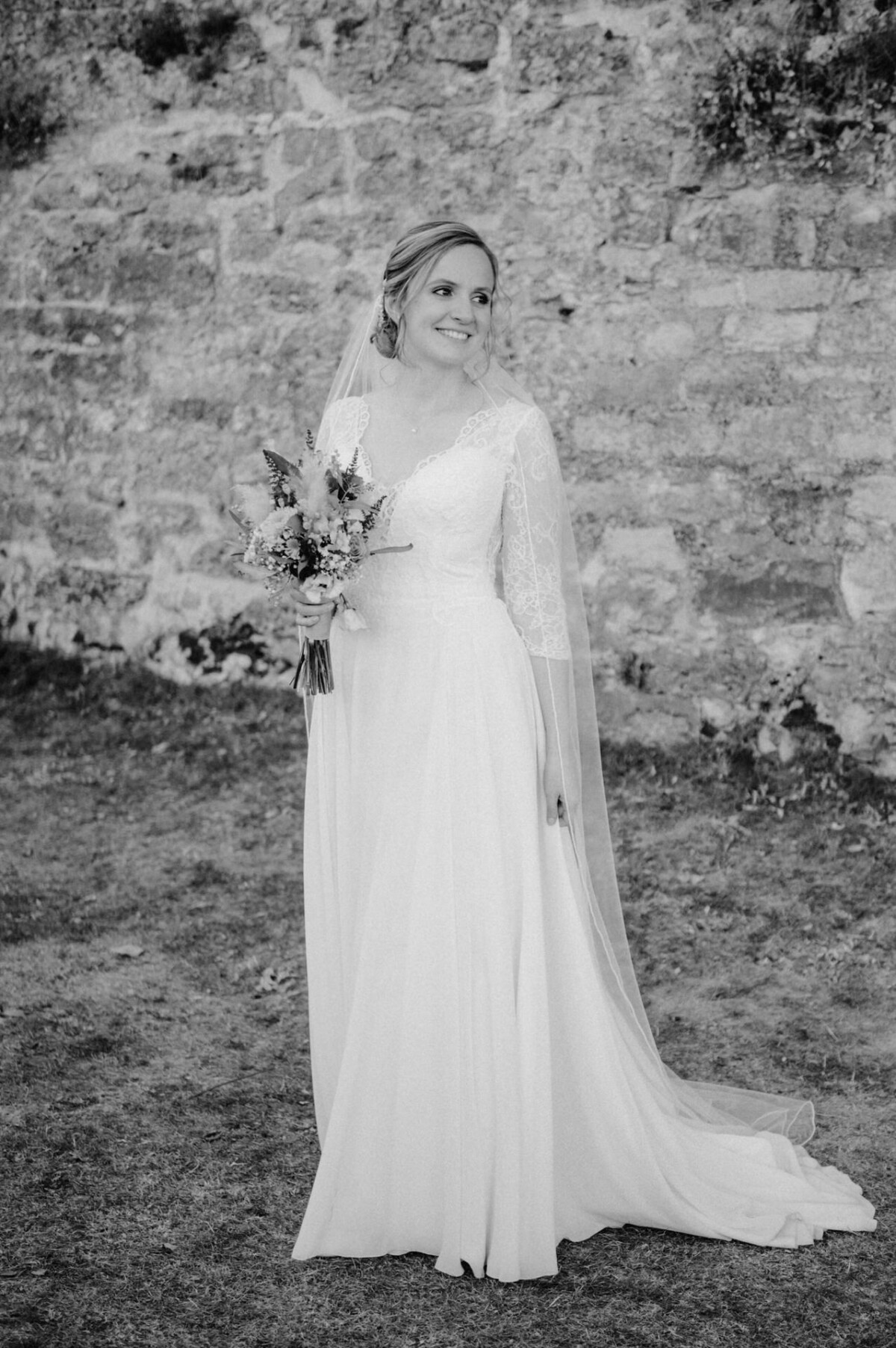 Eine Braut in einem Brautkleid mit Spitzenärmeln hält einen Blumenstrauß in der Hand, steht lächelnd auf dem Gras vor einer Steinmauer im Hintergrund.