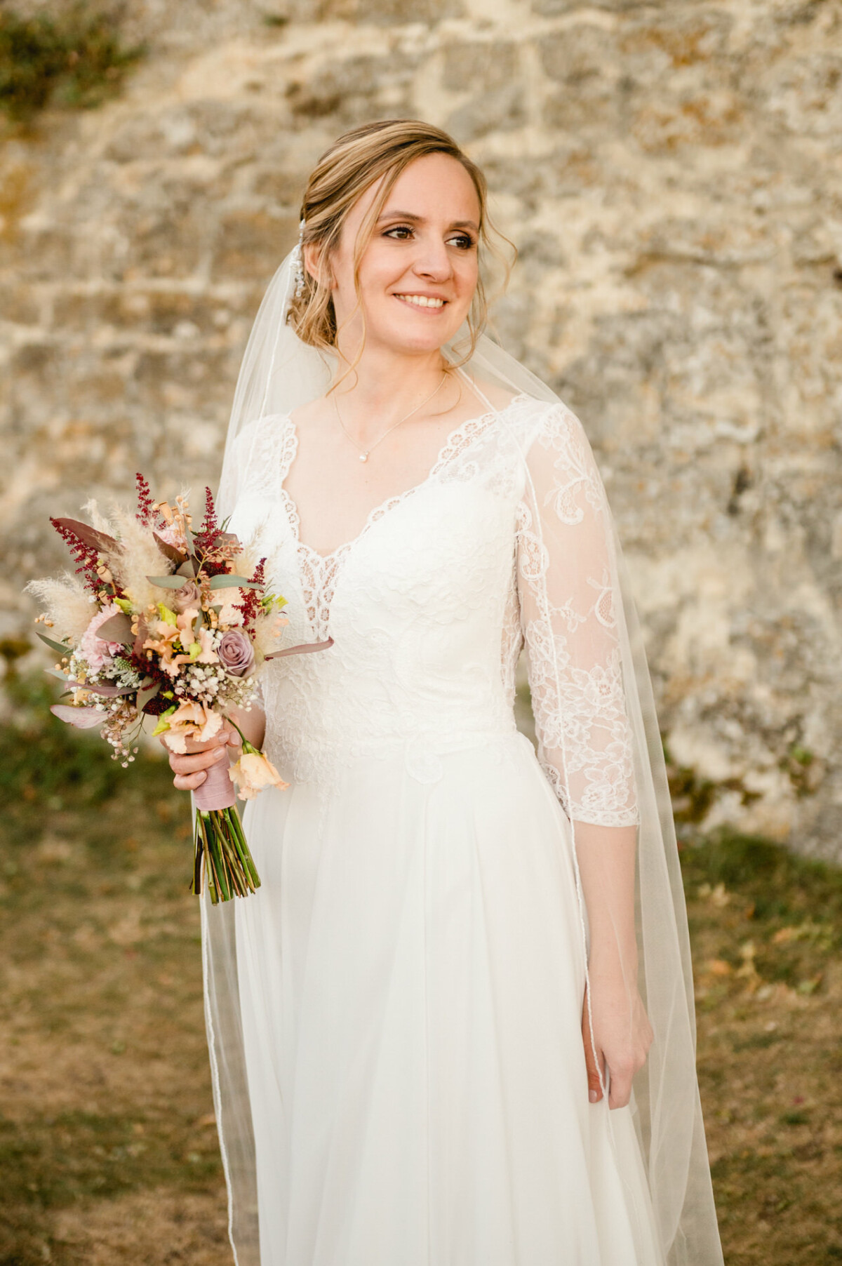 Eine Braut in einem Spitzenbrautkleid hält einen Blumenstrauß und steht im Freien mit einer Steinmauer im Hintergrund.
