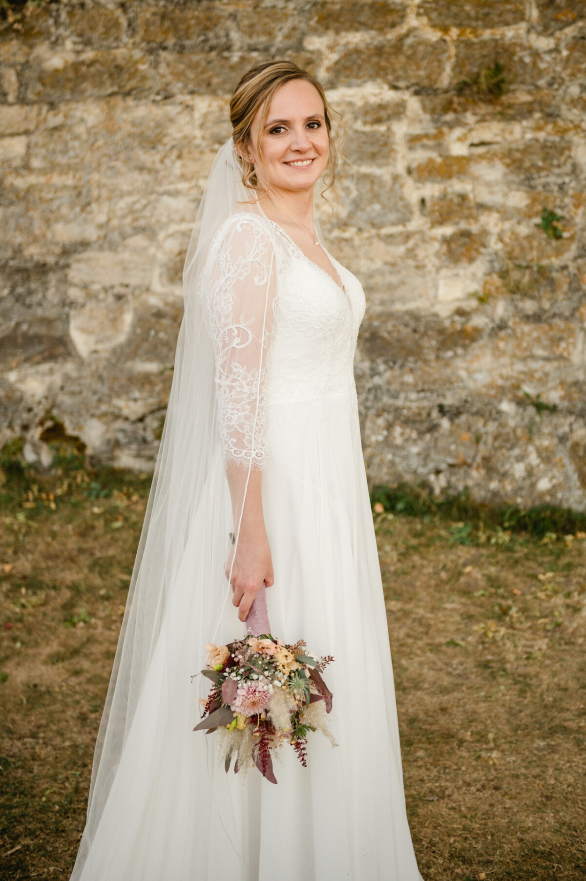 Eine Braut in einem langen weißen Kleid und Schleier hält einen Blumenstrauß und steht im Freien vor einer Steinmauer.