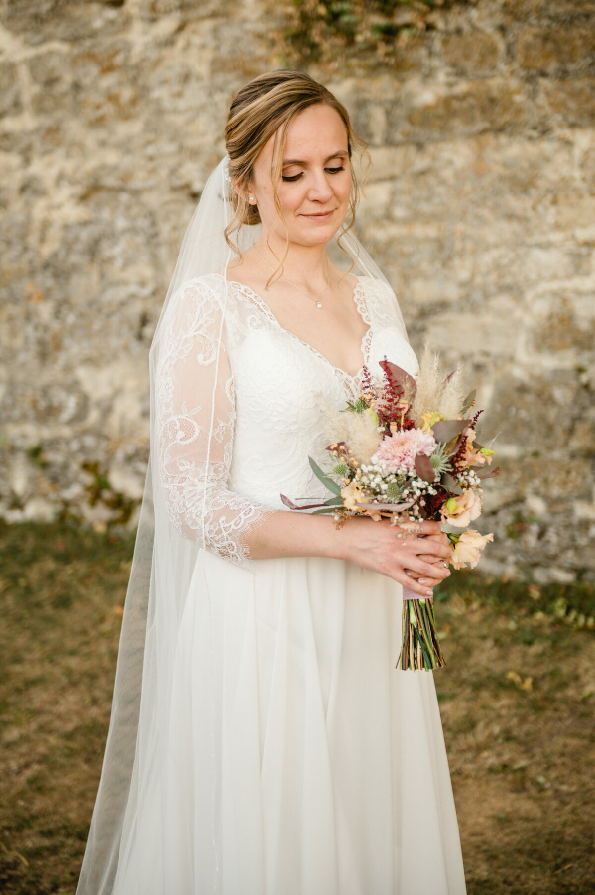 Eine Braut in einem weißen Spitzenkleid hält einen Blumenstrauß und steht vor einer Steinmauer.
