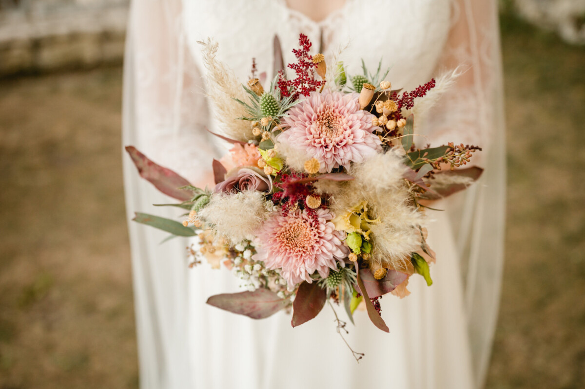 Eine Braut hält vor einem unscharfen Hintergrund im Freien einen Strauß mit rosa Blumen, grünen Disteln, braunen Blättern und kleinen weißen und gelben Blüten.
