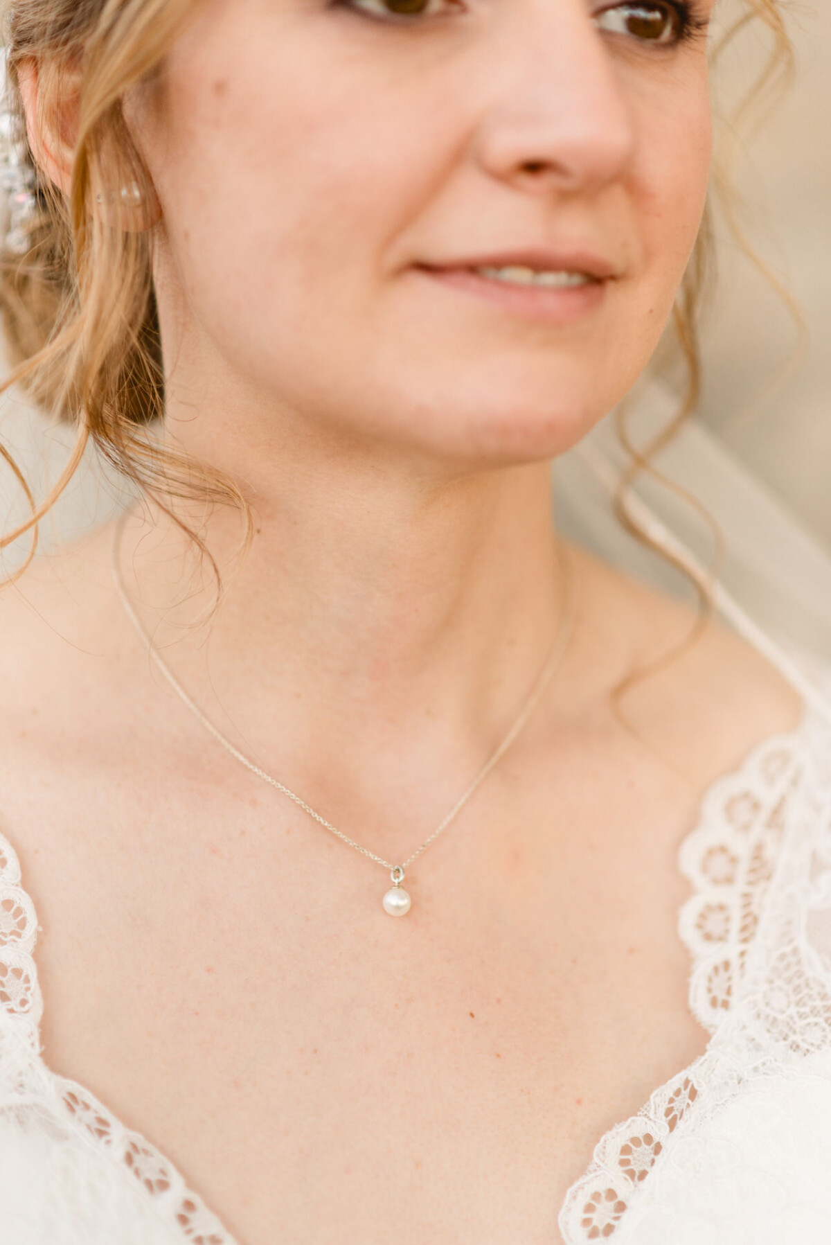 christian-lorenz.net Eine Frau in einem Spitzenbrautkleid trägt eine Perlenkette. Ihr Haar ist zu lockeren Locken frisiert, und sie lächelt leicht. Der Hintergrund ist leicht verschwommen.
