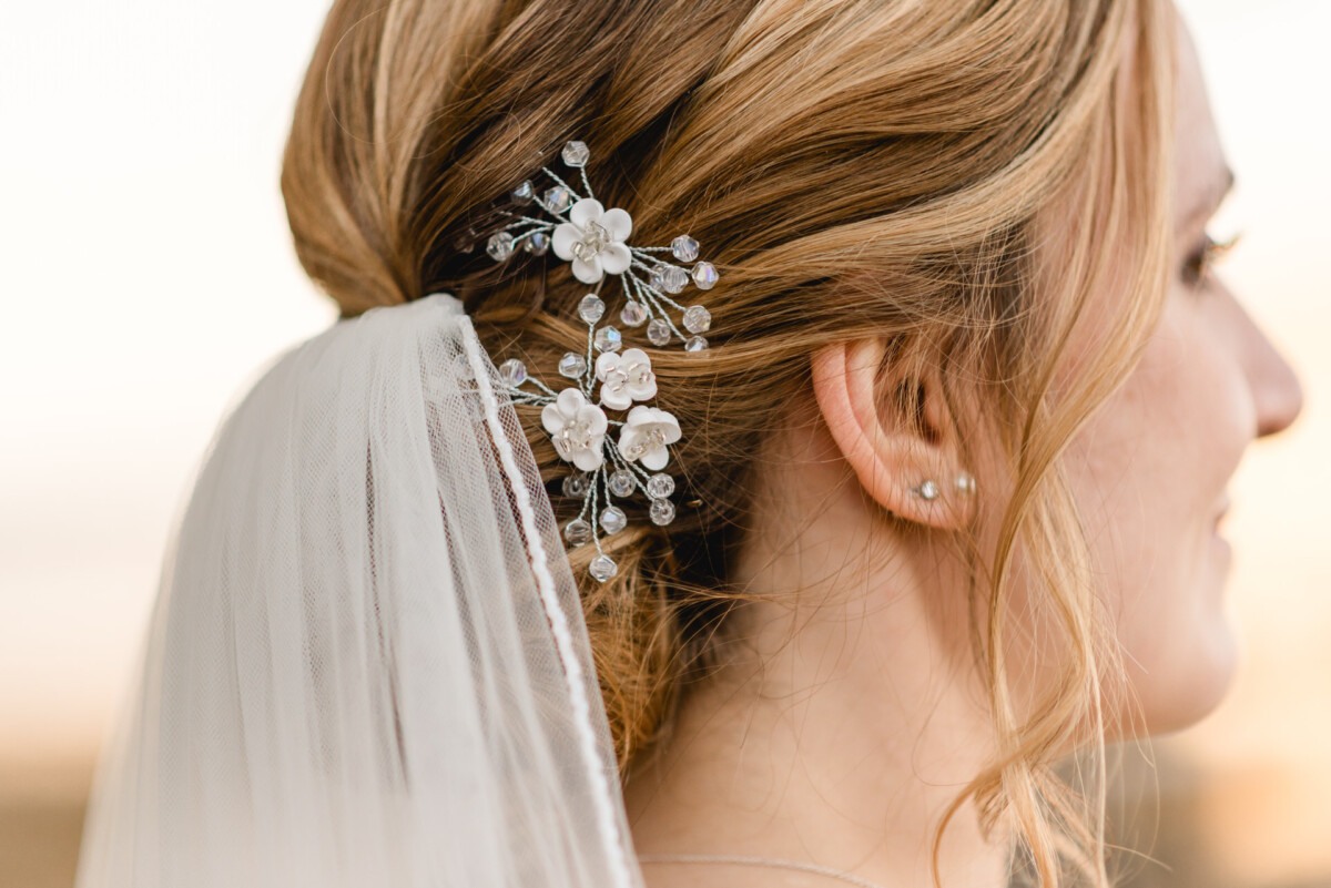 christian-lorenz.net Seitenprofil der Braut mit hochgestecktem Haar, geschmückt mit einem weißen Haarteil mit Blumenmuster und einem Schleier, vor einem unscharfen Hintergrund.