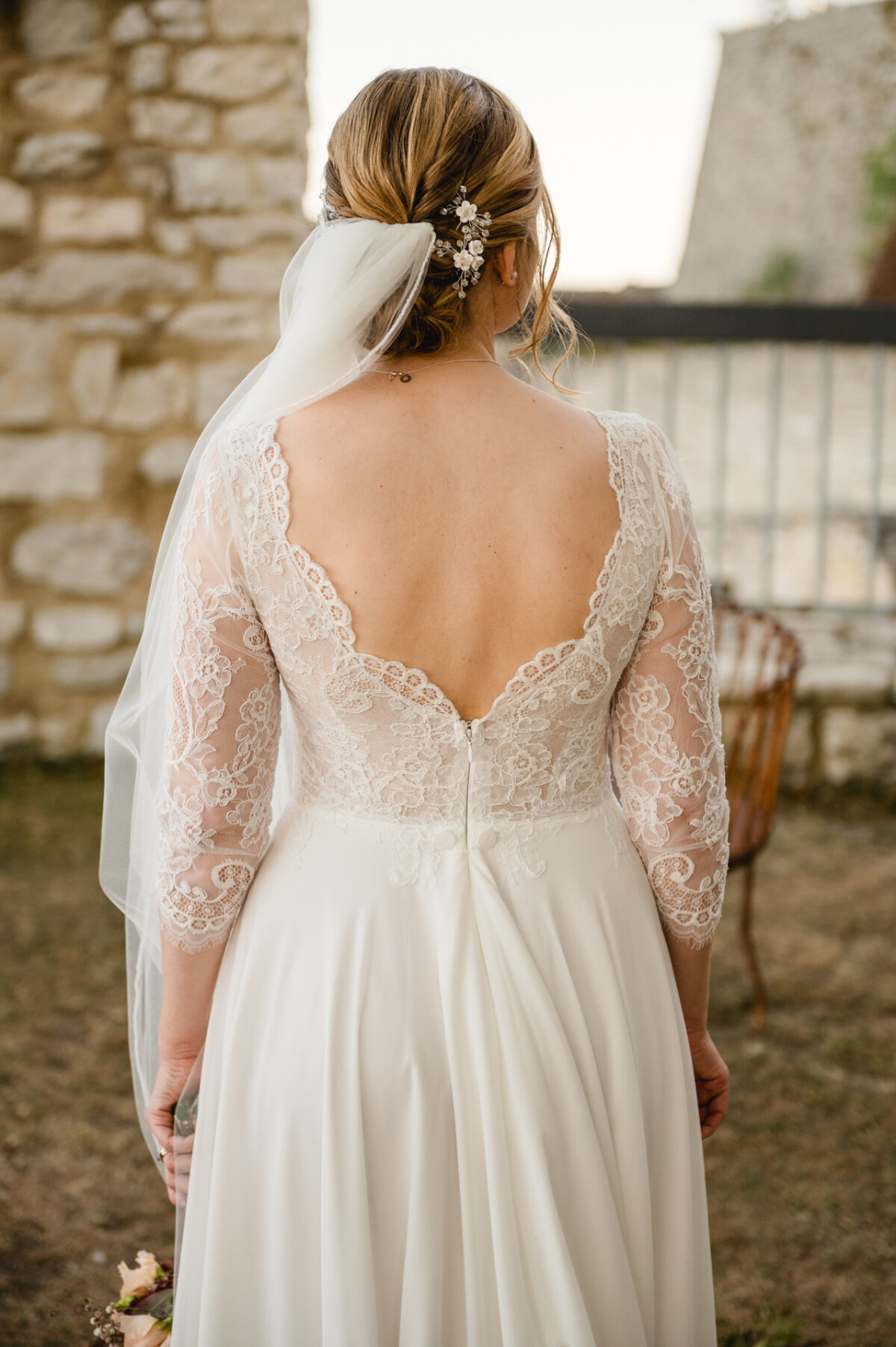 Braut in einem Spitzenbrautkleid mit tiefem Rückenausschnitt hält einen Blumenstrauß in der Hand. Ihr Haar ist hochgesteckt, mit dekorativer Spange und Schleier. Sie steht im Freien neben einer Steinmauer und einem Metallzaun.