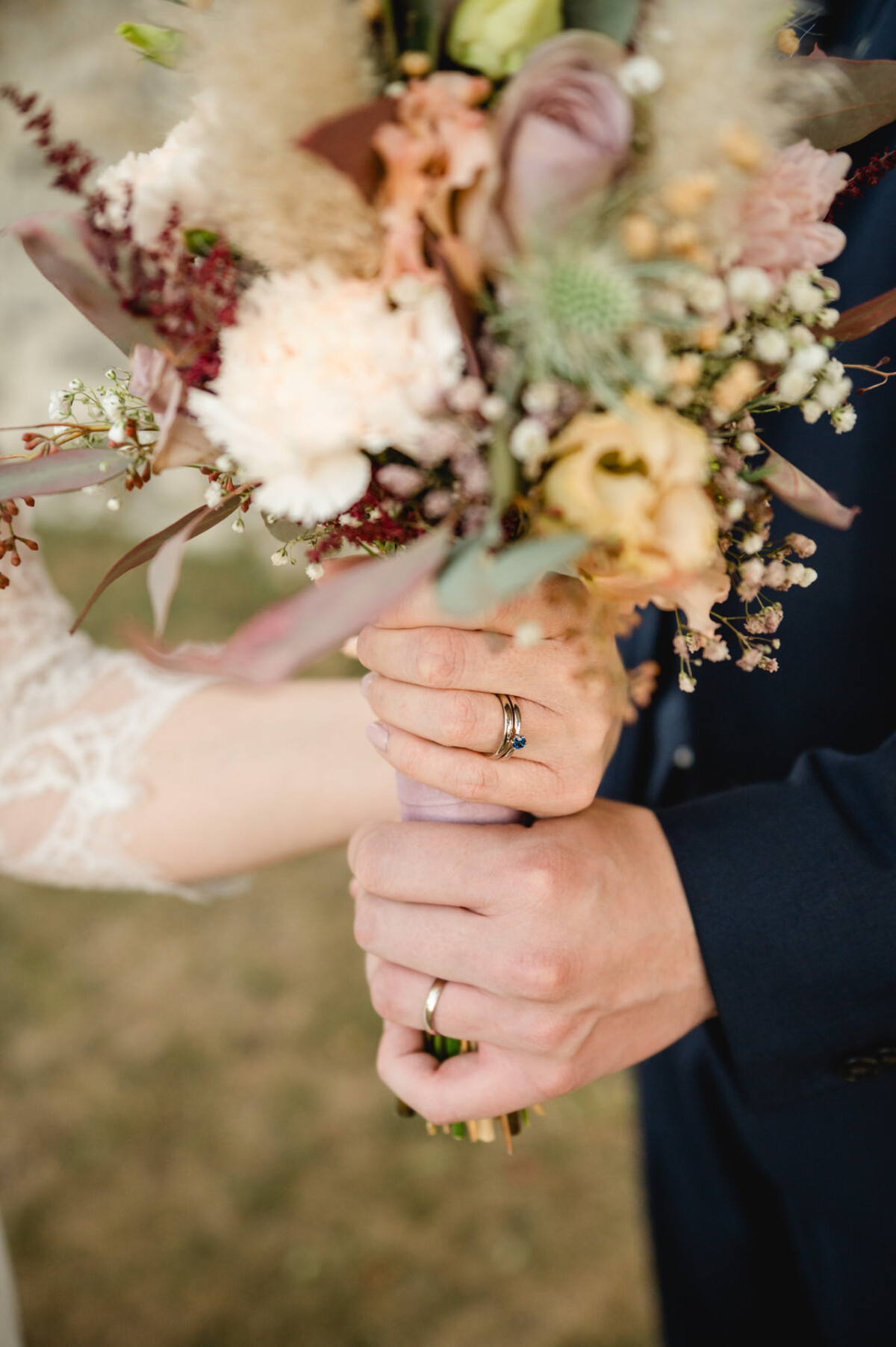 Nahaufnahme eines Paares mit einem Brautstrauß. Die Person im dunklen Anzug hält die Stiele, während die Person mit den Spitzenärmeln ihre Hand darauf legt. Beide tragen Eheringe.