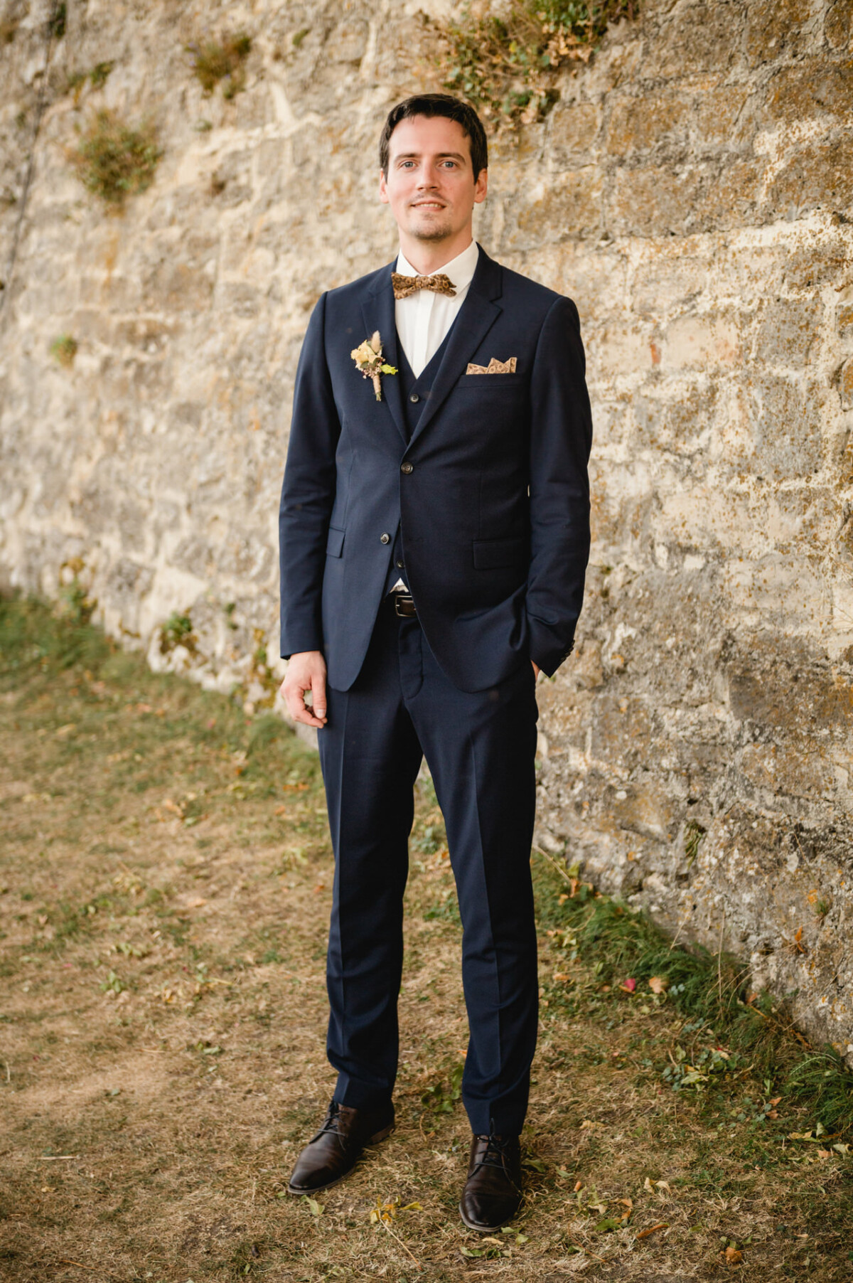 Ein Mann in einem dunklen Anzug mit Fliege steht auf dem Gras neben einer Steinmauer.