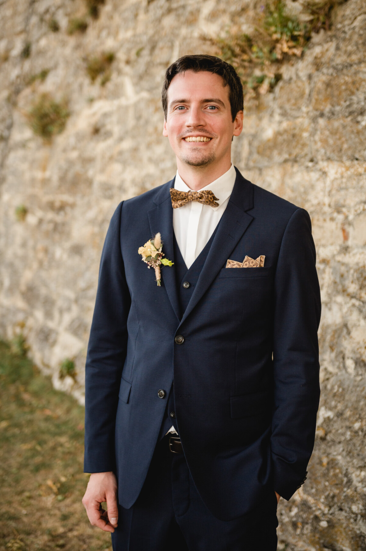 Ein Mann in einem dunklen Anzug mit Fliege lächelt, während er im Freien vor einer Steinmauer steht.