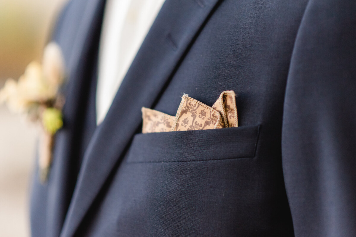 christian-lorenz.net Nahaufnahme eines marineblauen Anzugsjacketts mit einem gemusterten braunen Einstecktuch und einer Boutonniere am Revers.