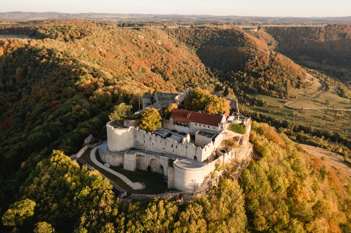 Luftaufnahme einer großen mittelalterlichen Burg, eingebettet in eine Waldlandschaft, umgeben von Hügeln mit Herbstlaub.