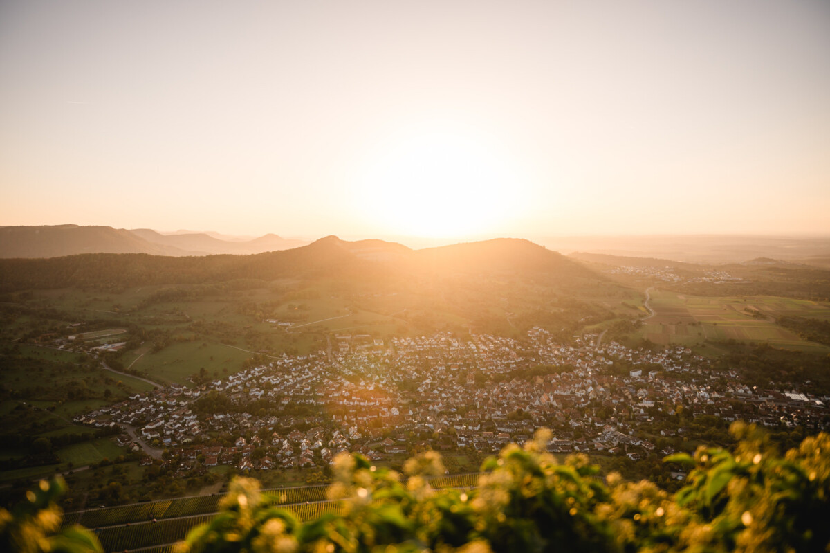 2024.10.05-Julia-Frank-Hochzeit-Burg-Hohen-Neuffen-bei-Metzingen-00695 Ein malerischer Blick auf eine Stadt mit verstreuten Häusern, umgeben von grünen Hügeln bei Sonnenuntergang. Die Sonne steht tief am Horizont und taucht die Landschaft in einen warmen Schein.