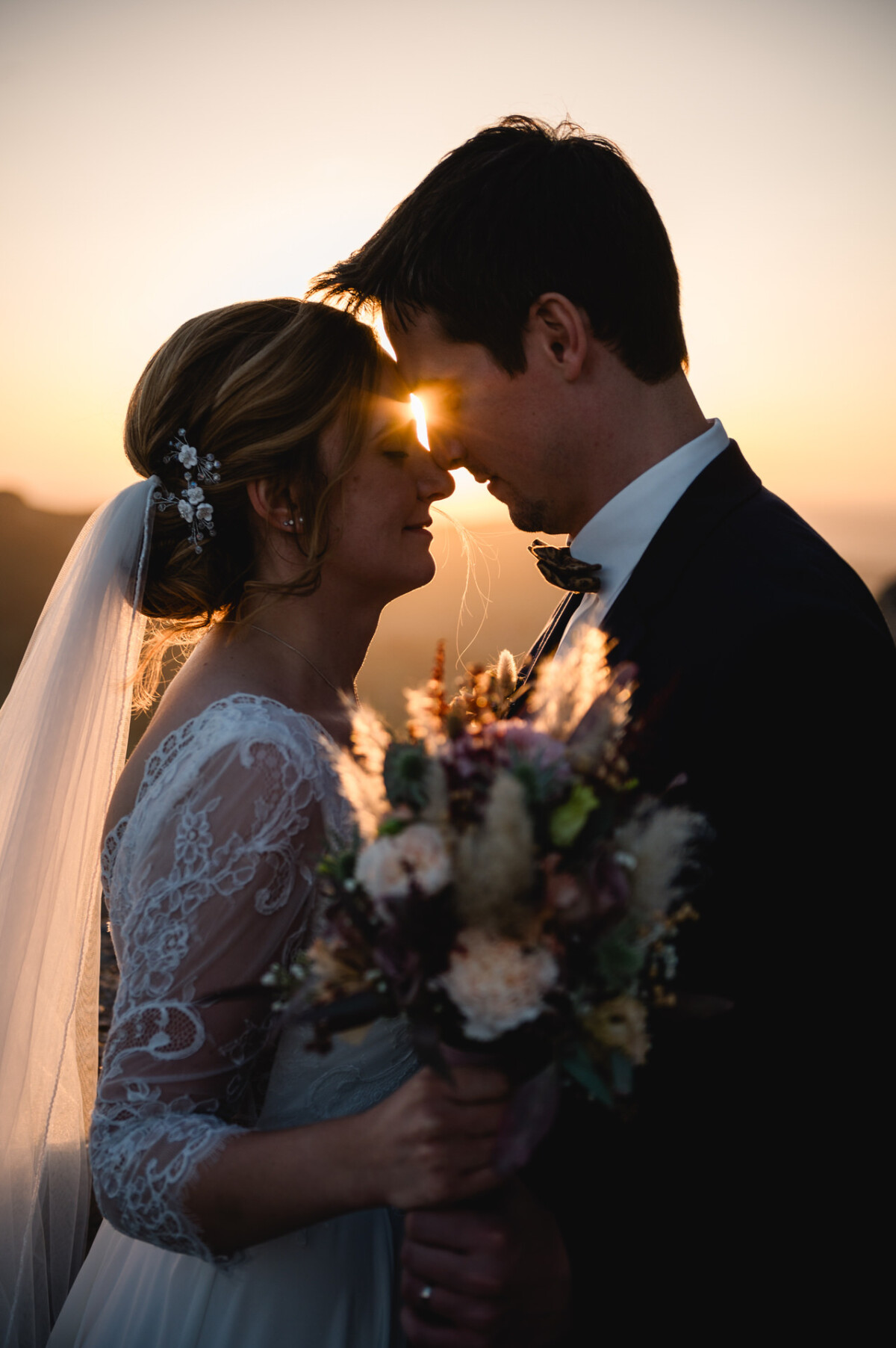 Braut und Bräutigam stehen bei Sonnenuntergang eng beieinander, berühren sich mit den Stirnen, sie hält einen Blumenstrauß.
