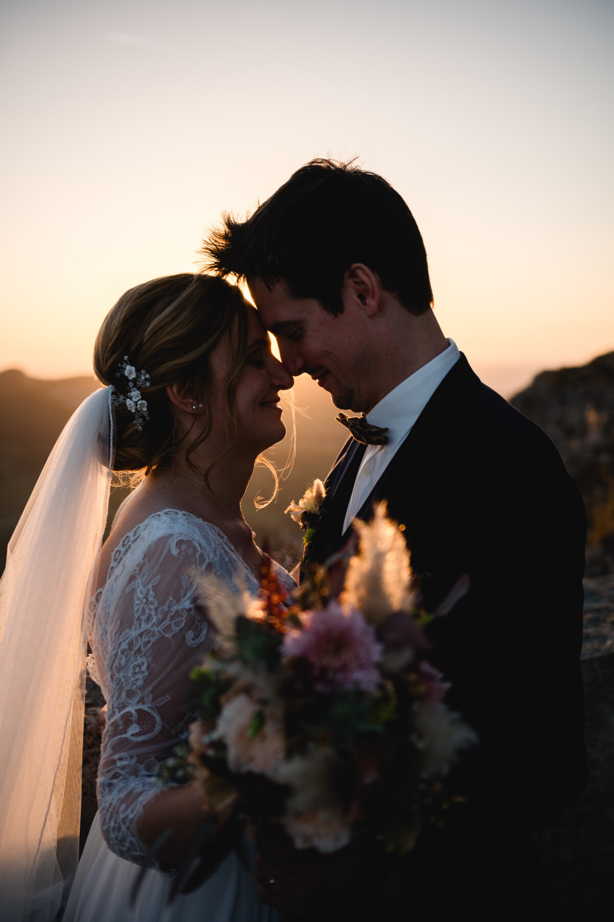 Braut und Bräutigam stehen bei Sonnenuntergang eng beieinander und berühren sich mit den Stirnen. Die Braut hält einen Blumenstrauß in der Hand und trägt ein Spitzenkleid mit Schleier; der Bräutigam trägt einen Anzug.