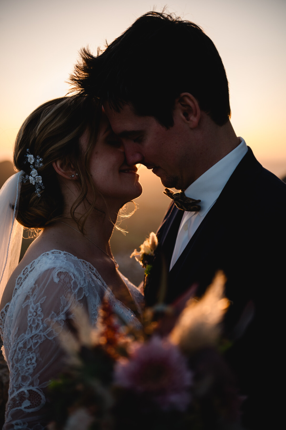 Braut und Bräutigam stehen sich bei Sonnenuntergang dicht gegenüber. Die Braut trägt einen Schleier und ein Spitzenkleid und hält einen Blumenstrauß in der Hand. Der Bräutigam trägt einen dunklen Anzug mit Fliege.