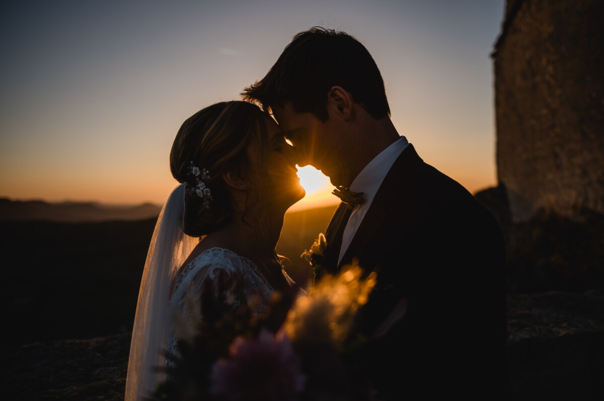 Ein Paar steht bei Sonnenuntergang eng beieinander und berührt seine Stirn. Die Braut trägt einen Schleier, der Bräutigam einen Anzug. Sanftes Licht erzeugt einen Silhouetteneffekt.