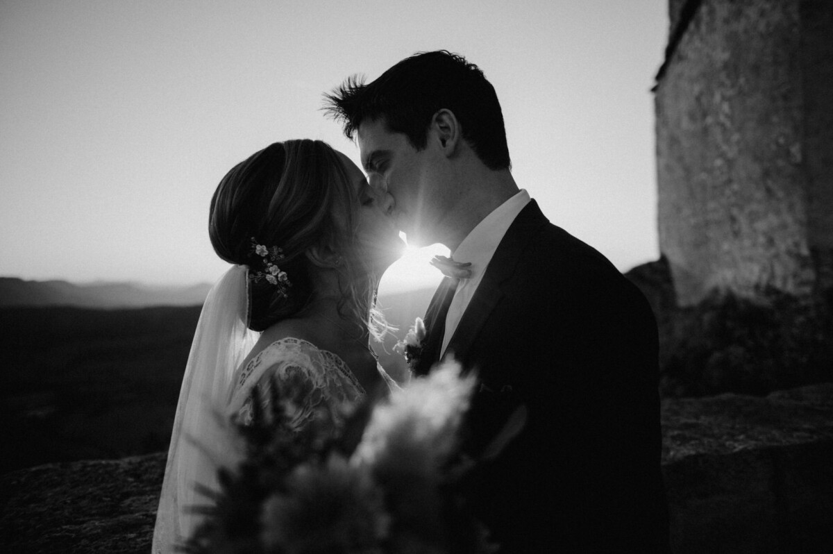 Braut und Bräutigam küssen sich bei Sonnenuntergang, wobei die Sonne einen Silhouetteneffekt erzeugt. Die Braut hält einen Blumenstrauß in der Hand. Beide sind in festlicher Kleidung und stehen neben einem Steingebäude.