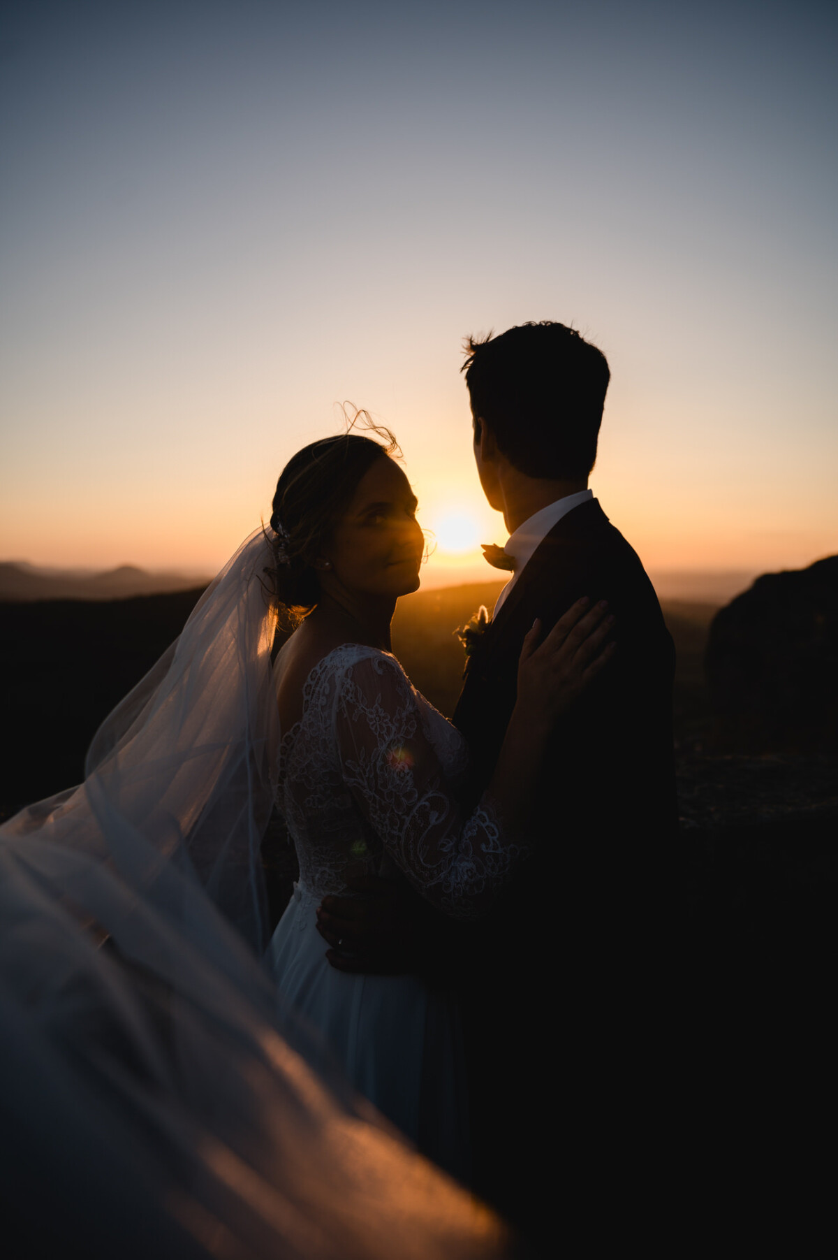 Braut und Bräutigam umarmen sich, die Silhouette zeichnet sich vor dem Hintergrund eines Sonnenuntergangs ab, wobei der Schleier der Braut sanft das Licht einfängt.