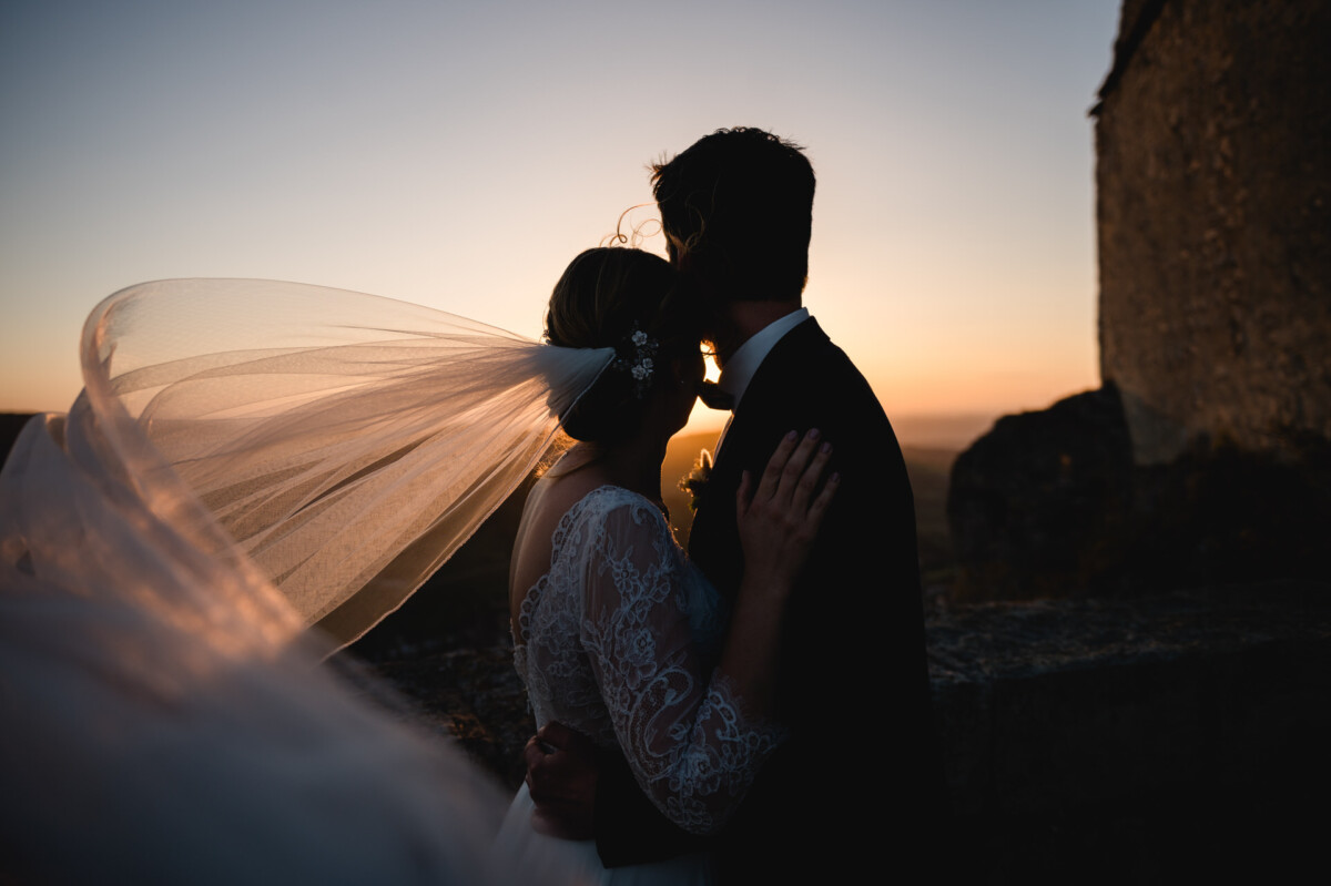 2024.10.05-Julia-Frank-Hochzeit-Burg-Hohen-Neuffen-bei-Metzingen-00716 Die Silhouette eines Paares umarmt sich bei Sonnenuntergang, der Schleier der Braut weht im Wind.