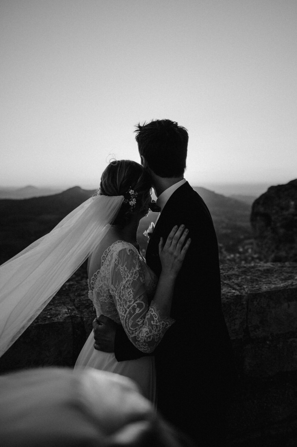 Braut und Bräutigam umarmen sich vor einer Berglandschaft bei Sonnenuntergang. Die Braut trägt einen Schleier und ein Spitzenkleid, der Bräutigam einen Anzug. Die Szene ist in Schwarzweiß gehalten.
