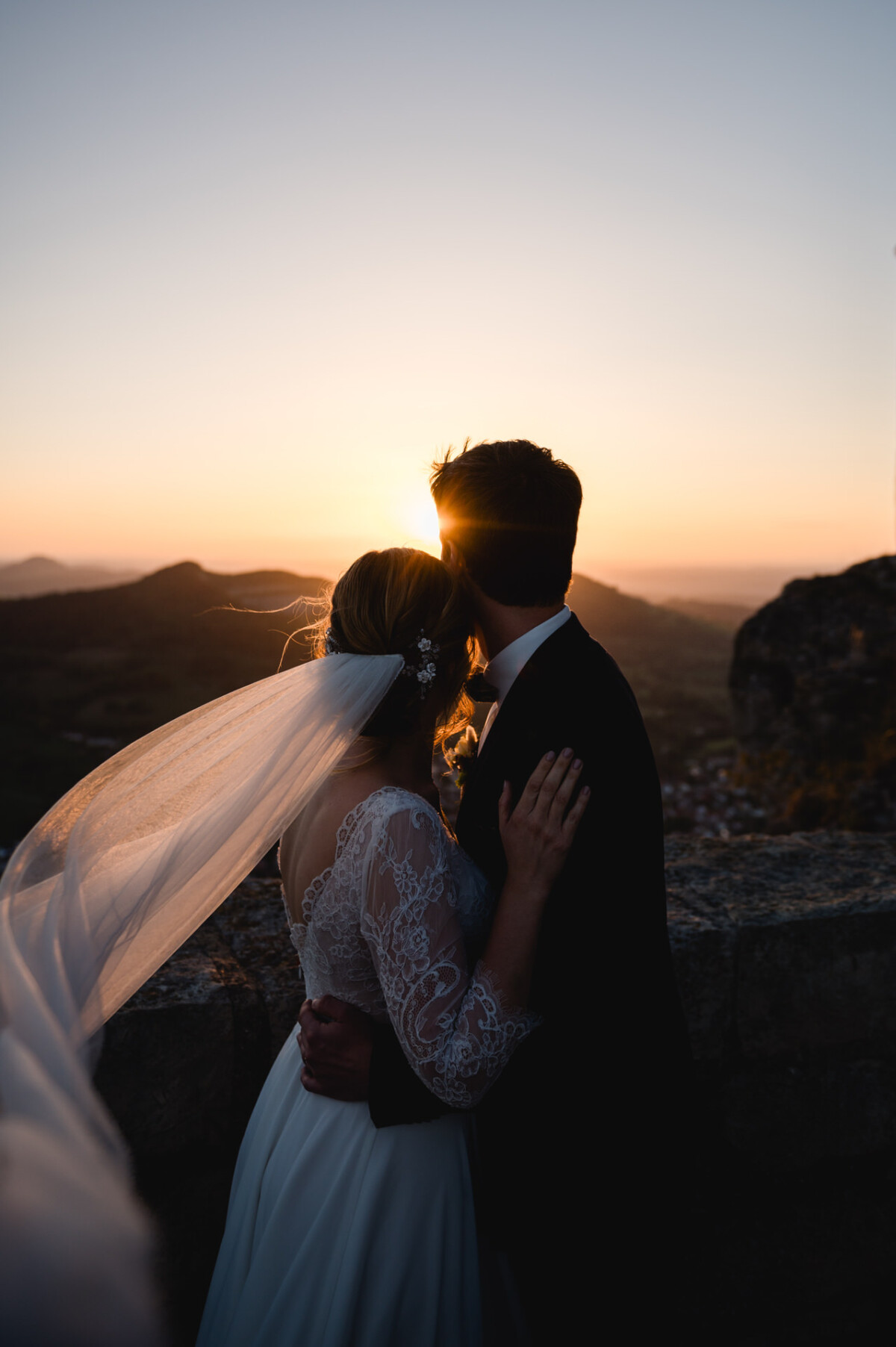 Ein Paar umarmt sich bei Sonnenuntergang auf einem Berg. Die Sonne geht hinter ihnen unter und wirft einen goldenen Schein auf die Landschaft. Der Schleier der Braut weht im Wind.