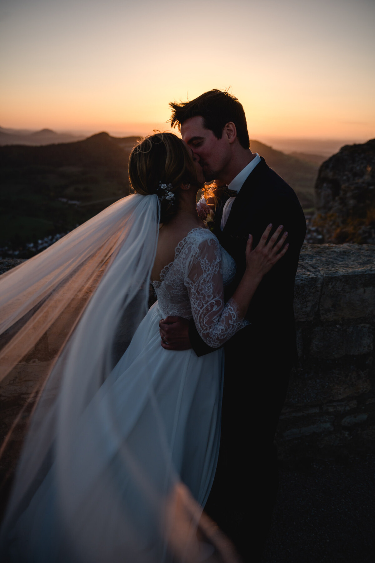 Ein Paar im Hochzeitskleid umarmt und küsst sich bei Sonnenuntergang, im Hintergrund eine Berglandschaft. Der Schleier der Braut weht im Wind.