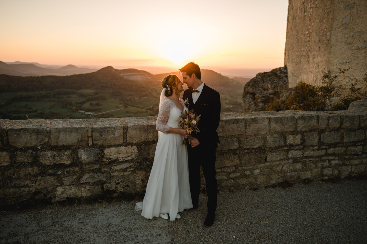 Braut und Bräutigam stehen dicht beieinander an einer Steinmauer, im Hintergrund ist ein malerischer Sonnenuntergang über fernen Hügeln zu sehen. Die Braut hält einen Blumenstrauß.
