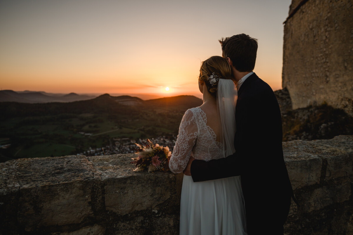 Ein Paar in Hochzeitskleidung umarmt sich und betrachtet von einem Steinbalkon mit Blick auf ein Tal den Sonnenuntergang.