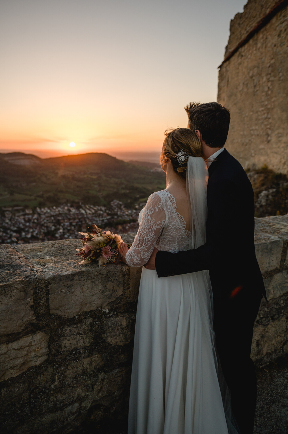 Braut und Bräutigam stehen auf einer Steinmauer und genießen den malerischen Sonnenuntergang mit Hügeln und einer entfernten Stadt. Die Braut hält einen Blumenstrauß in der Hand und trägt einen langen Schleier.