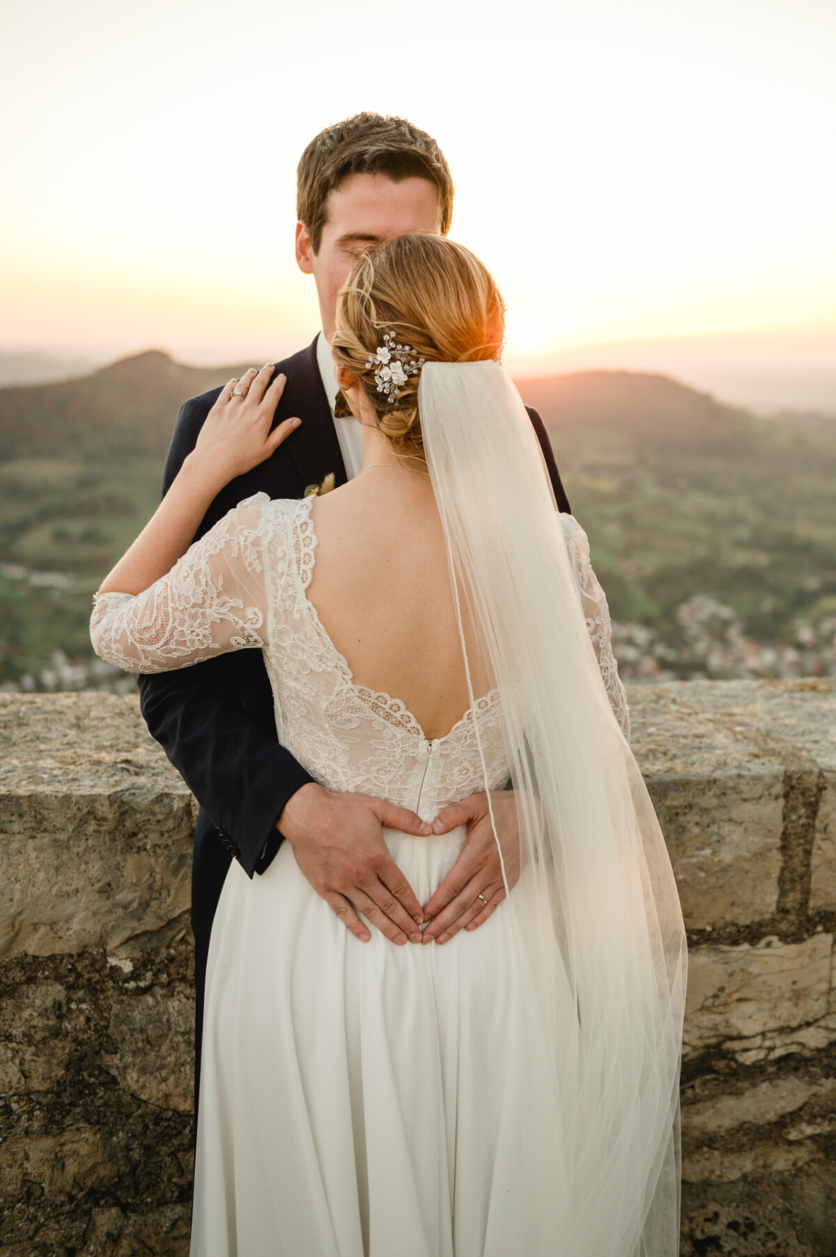 Ein Paar umarmt sich im Hochzeitskleid und steht einander mit den Händen auf der Hüfte gegenüber. Im Hintergrund ist ein malerischer Sonnenuntergang über einer ländlichen Landschaft zu sehen.