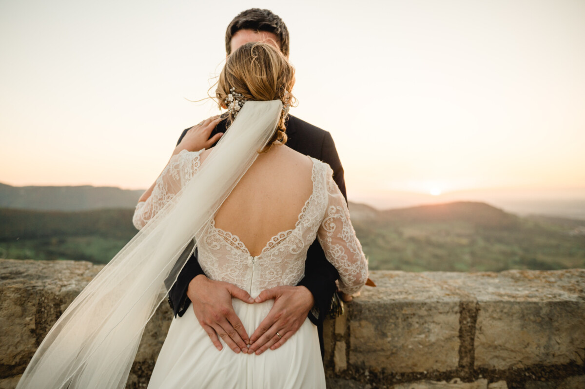 Ein Paar in Hochzeitskleidung umarmt sich bei Sonnenuntergang an einer Steinmauer, wobei die Braut der Kamera den Rücken zuwendet und einen langen Schleier trägt.