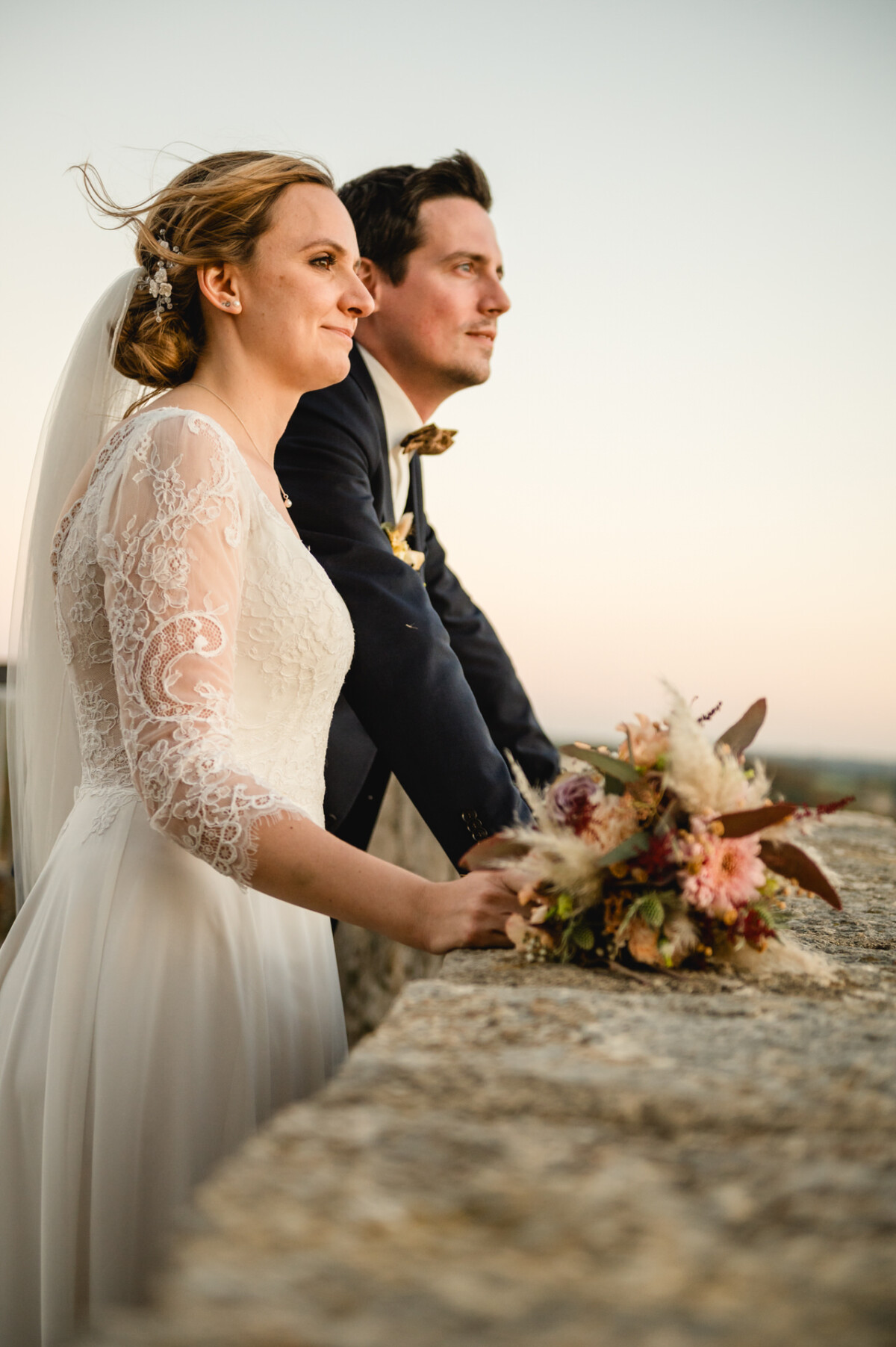 Braut und Bräutigam stehen bei Sonnenuntergang gemeinsam im Freien und blicken in die Ferne. Die Braut hält einen Blumenstrauß in der Hand.