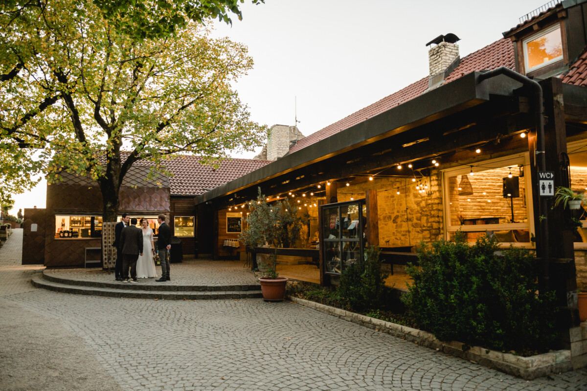 Eine Außenterrasse mit Lichterketten und Backsteingebäuden. Eine Gruppe von Menschen unterhält sich neben einem großen Baum mit grünen Blättern. Der Boden ist mit Kopfsteinpflaster gepflastert.