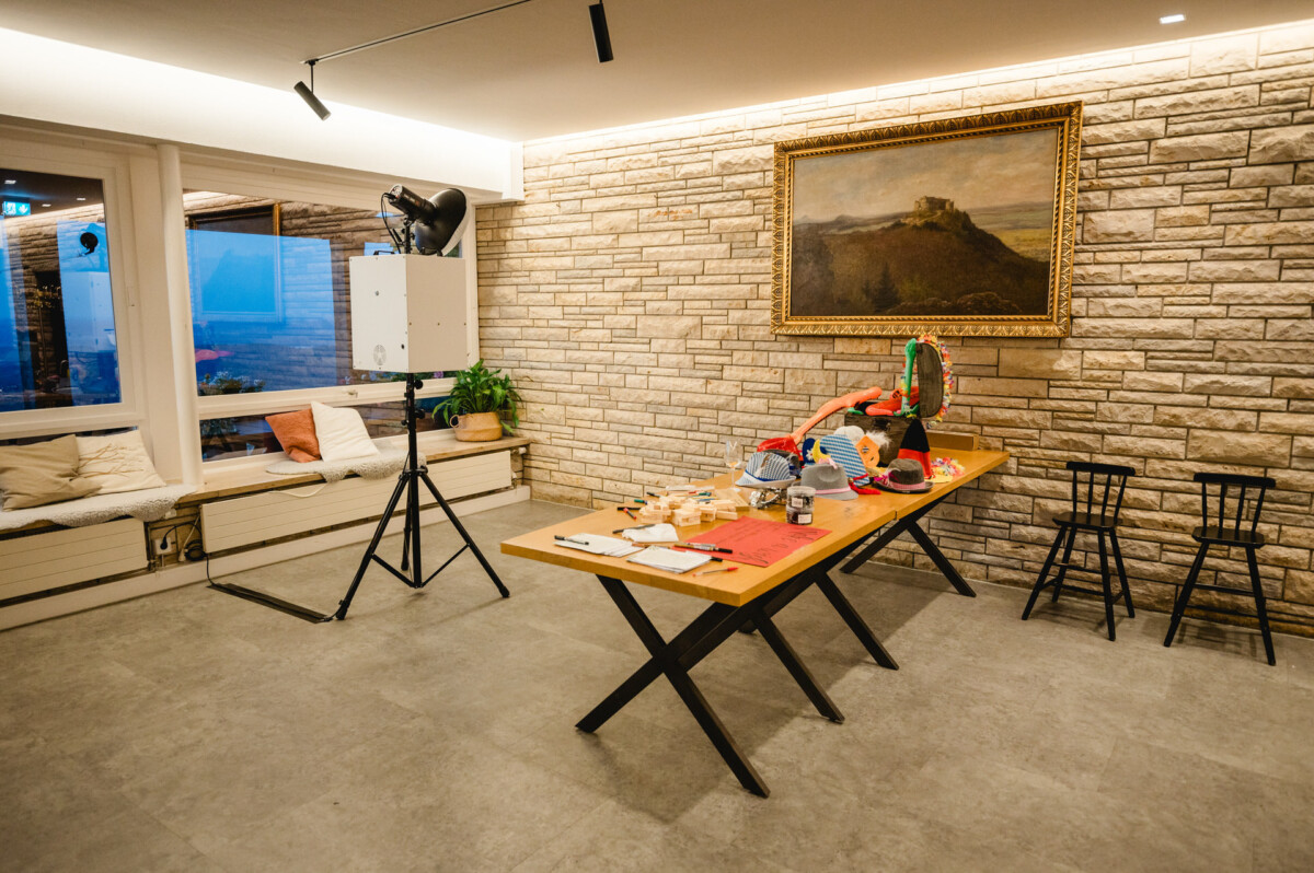 Raum mit Steinwänden, einem Tisch mit Werkzeugen und einem roten Gegenstand sowie zwei schwarzen Stühlen. An der Wand hängt ein Gemälde, und unter heller Deckenbeleuchtung ist ein Fensterplatz sichtbar.