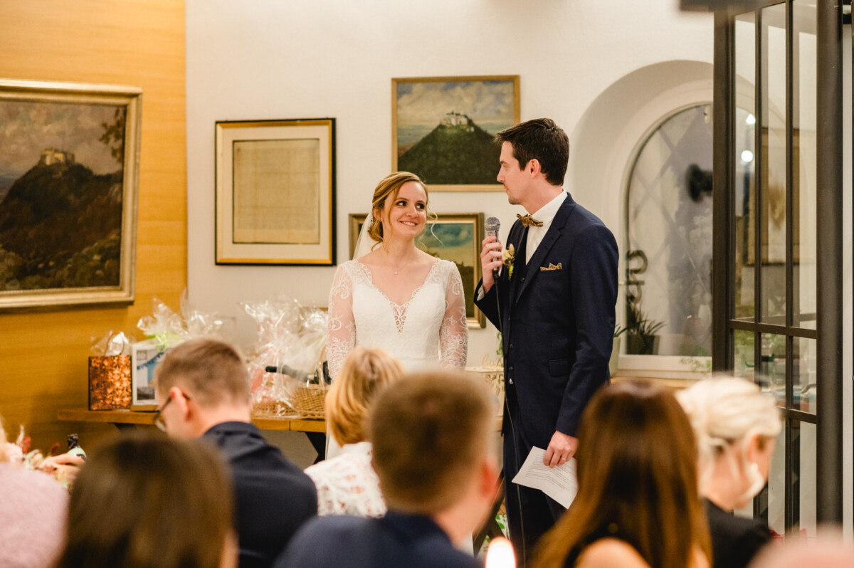 Braut und Bräutigam stehen in einem Raum. Der Bräutigam hält ein Mikrofon in der Hand. Um sie herum sitzen Gäste. An der Wand hinter ihnen hängen verschiedene gerahmte Kunstwerke.