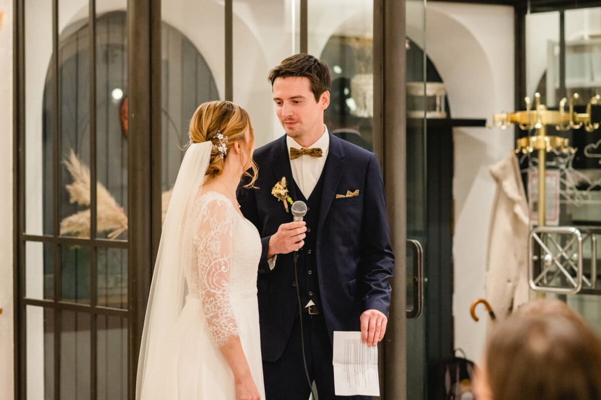 Braut und Bräutigam stehen im Innenbereich, der Bräutigam hält ein Mikrofon und ein Papier in der Hand und blickt einander an. Die Braut trägt ein weißes Kleid und ein Haarteil; der Bräutigam trägt einen dunklen Anzug und eine Fliege.