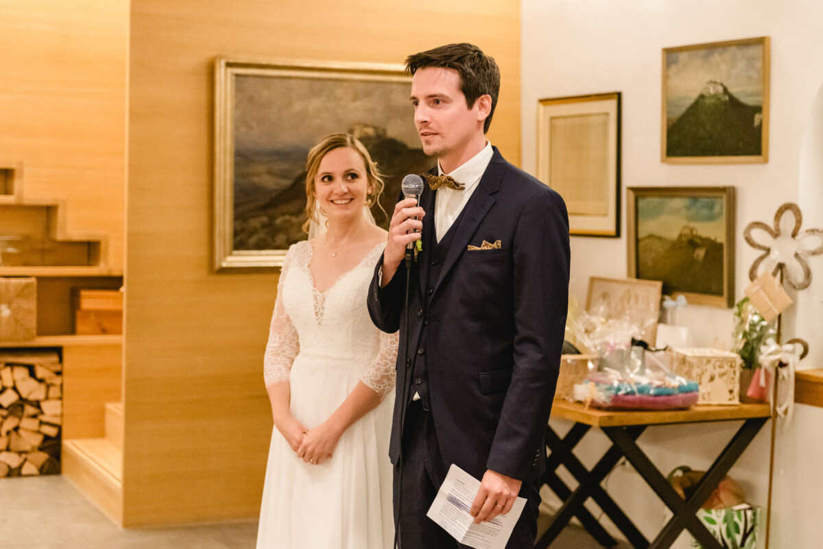 christian-lorenz.net Ein Mann im Anzug spricht in ein Mikrofon und hält dabei ein Blatt Papier in der Hand. Eine Frau in einem weißen Kleid steht lächelnd neben ihm. Sie befinden sich in einem Gebäude, hinter dem gerahmte Kunstwerke hängen.