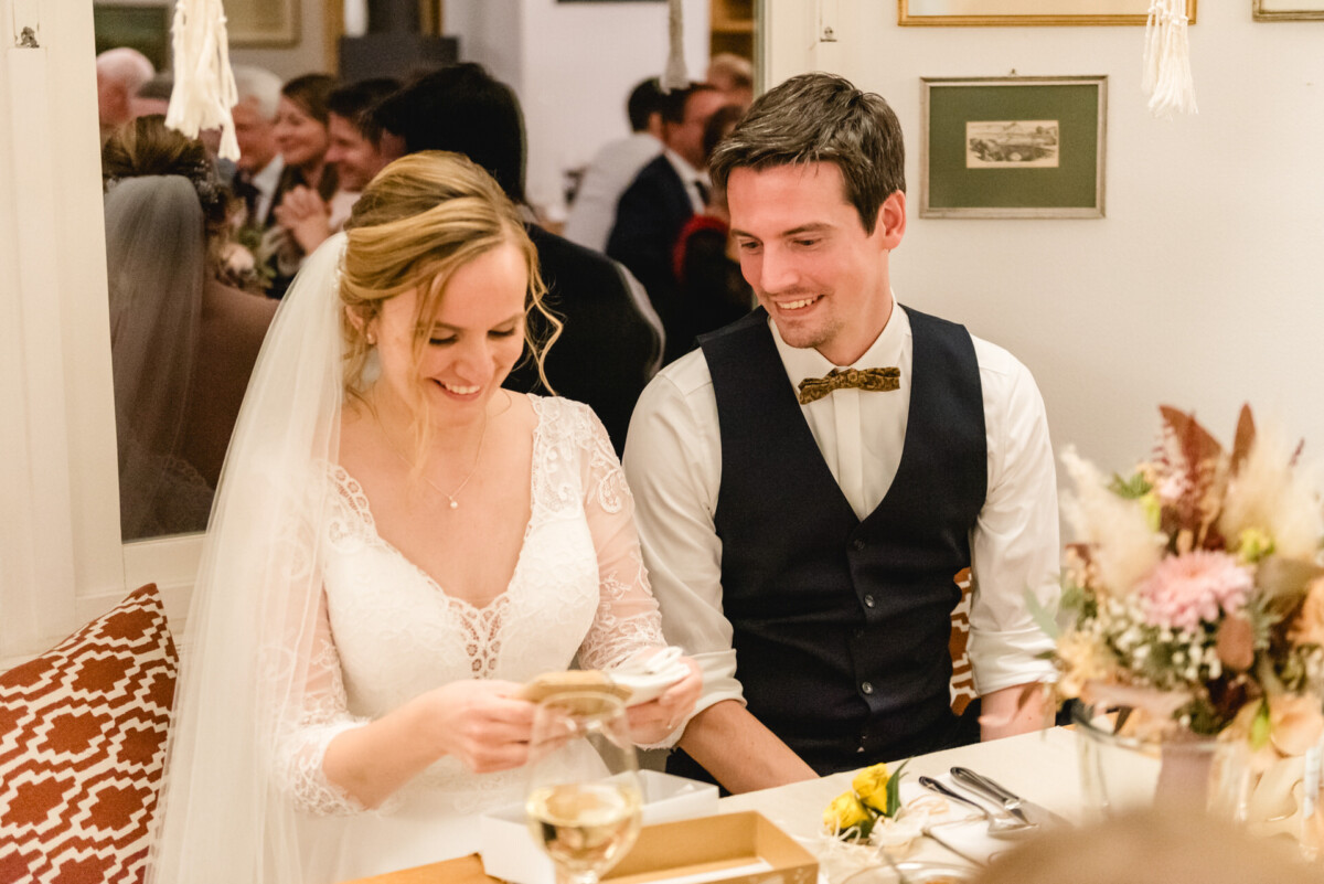 christian-lorenz.net Die Braut im weißen Kleid liest eine Karte, während der Bräutigam in Weste und Fliege lächelt neben ihr an einem Tisch mit Blumen und einem Weinglas.