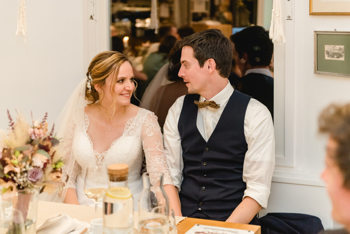 christian-lorenz.net Braut und Bräutigam sitzen lächelnd zusammen an einem dekorierten Hochzeitsempfangstisch.